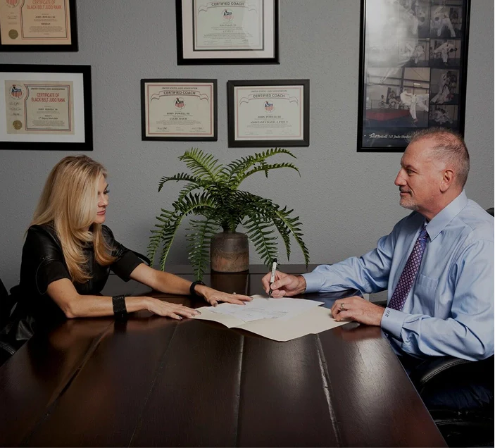 Attorney John Powell and his wife Stacia sitting at a table reviewing documents in an office with framed certificates and a potted plant on the wall.