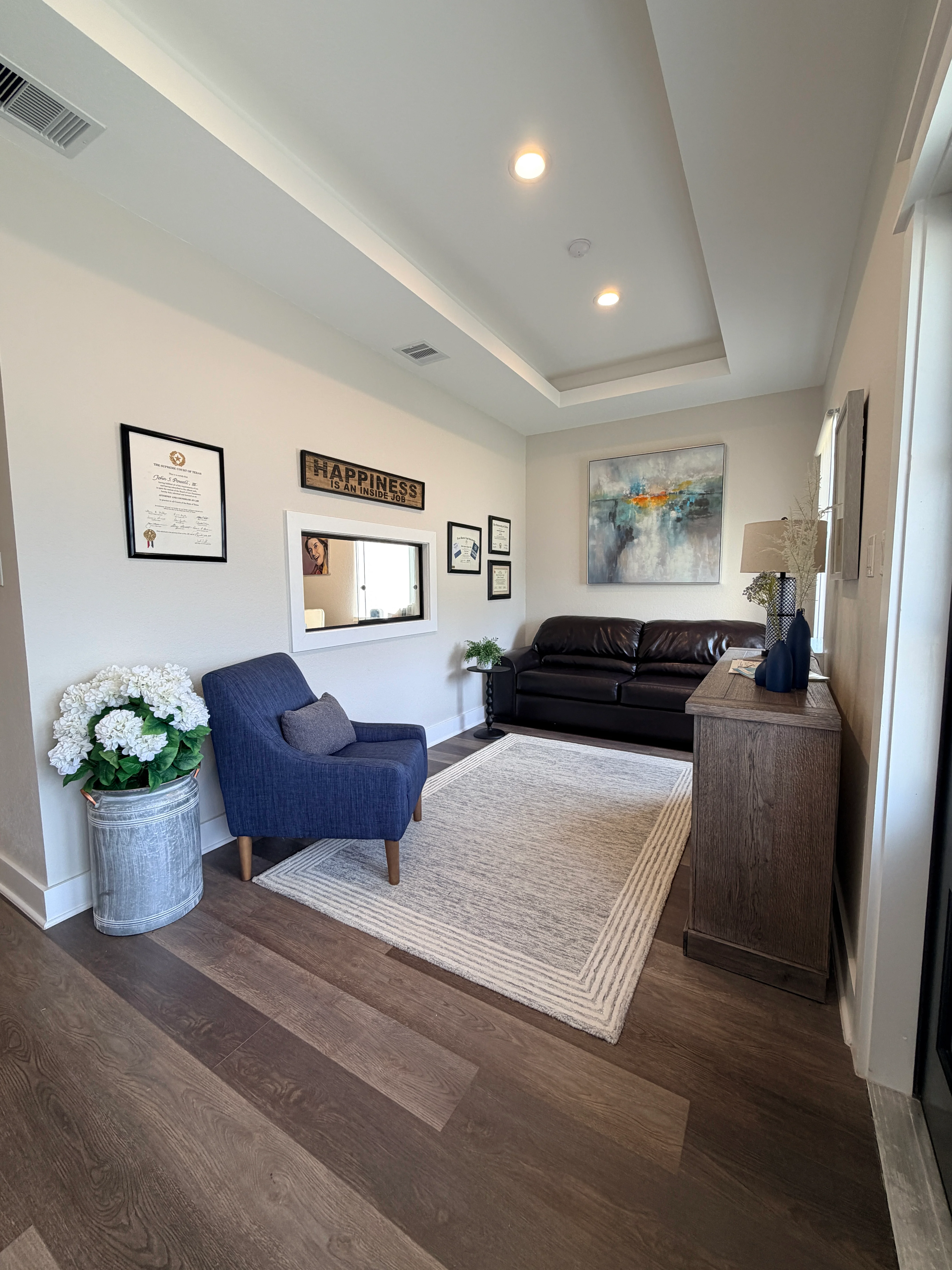 Modern law office waiting area with a blue chair, leather sofa, framed certificates, artwork, and decorative furnishings.