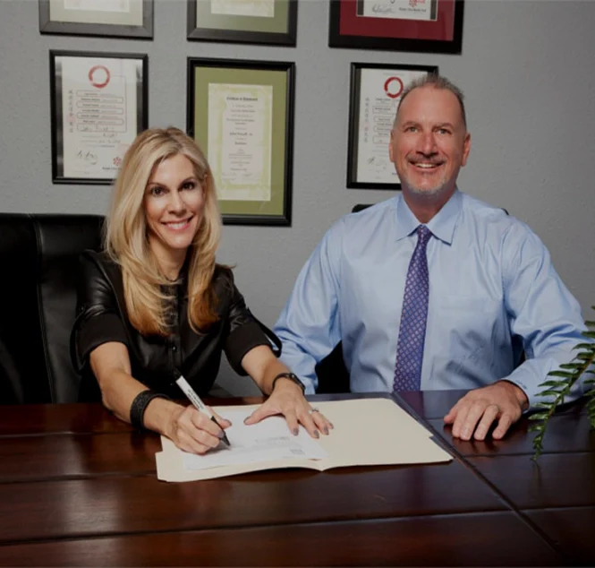 Attorney John Powell and his wife Stacia sit at a desk with framed certificates on the wall, one signing a document while the other looks on.